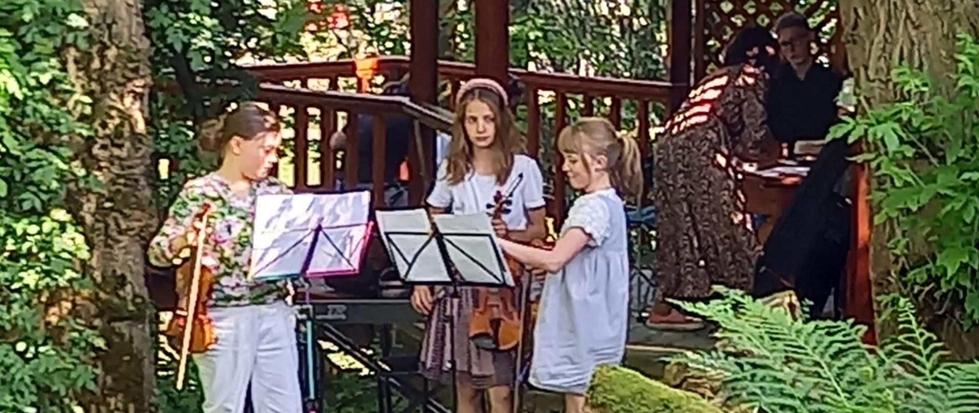 Zdjęcie w ogrodzie. Na środku gra trio skrzypcowe spoglądając na nuty umieszczone na dwóch pulpitach, za nimi keybord. Następnie między drzewami drewniana altanka, w której są uczestnicy koncertu