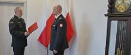 Strażak w mundurze wyjściowym trzyma czerwoną teczkę przed nim stoi funkcjonariusz Państwowej Straży Pożarnej w mundurze wyjściowym obok stoją dwie flagi Polski oraz zegar.