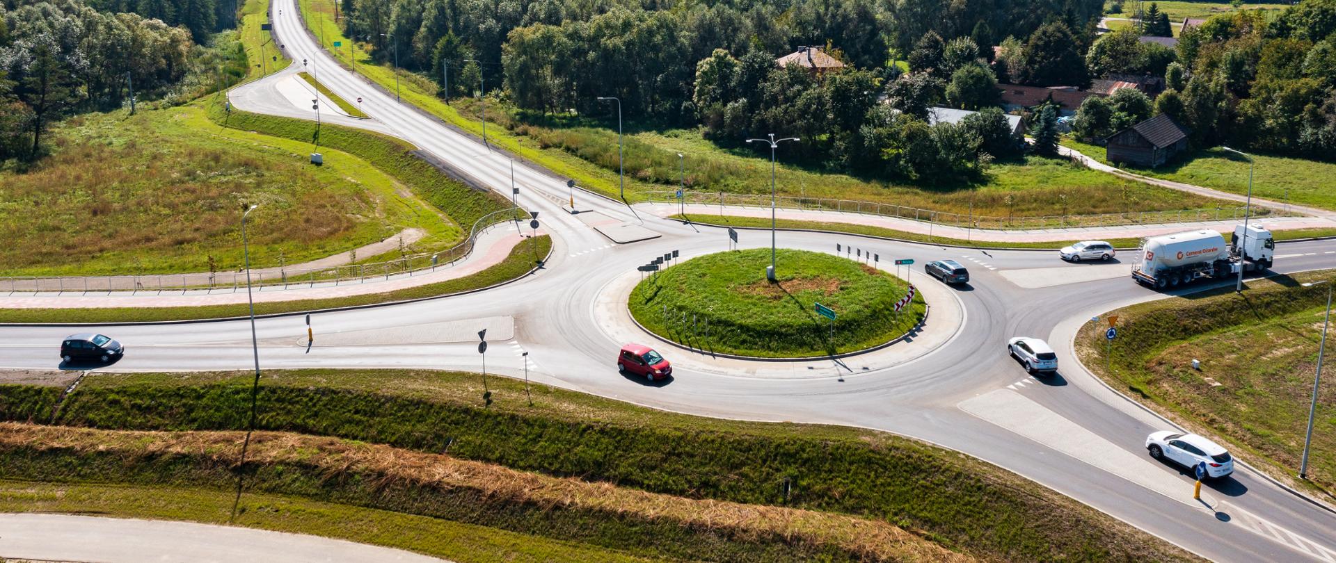 Widok z góry na rondo na obwodnicy Stalowej Woli i Niska. PO rondzie i na dojazdach poruszają się auta osobowe i ciężarowe. Świeci słońce, aura letnia. 