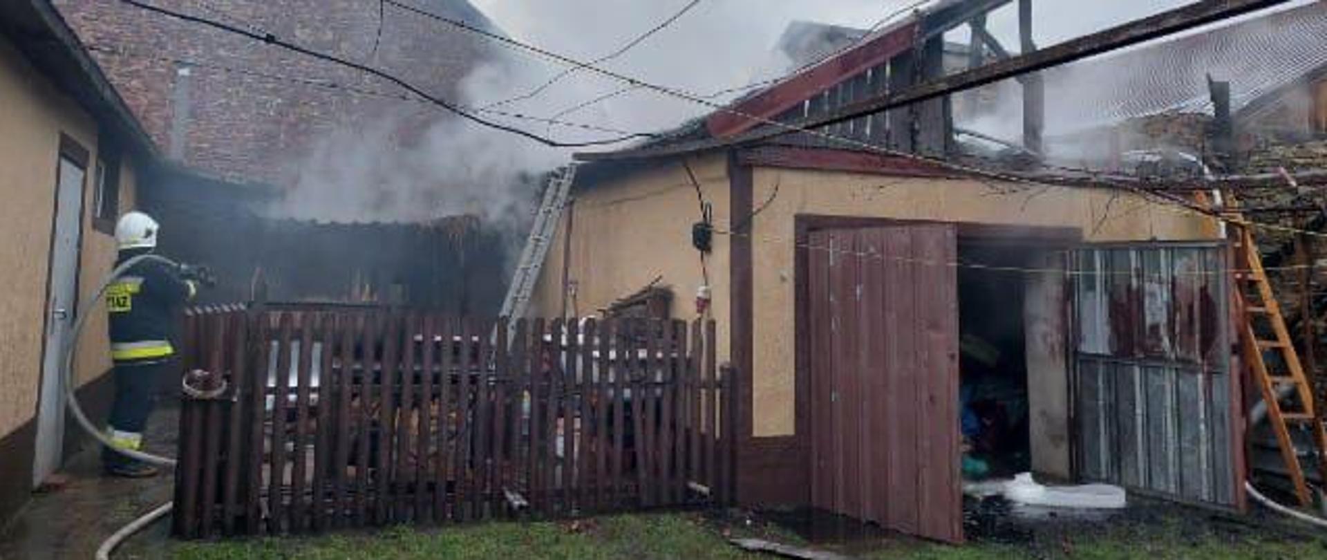 Na zdjęciu po lewej stronie stoi spalony garaż, centralnie i z lewej widać zagrożone budynki, za płotem stoi strażak z wężem gaśniczym na ramieniu