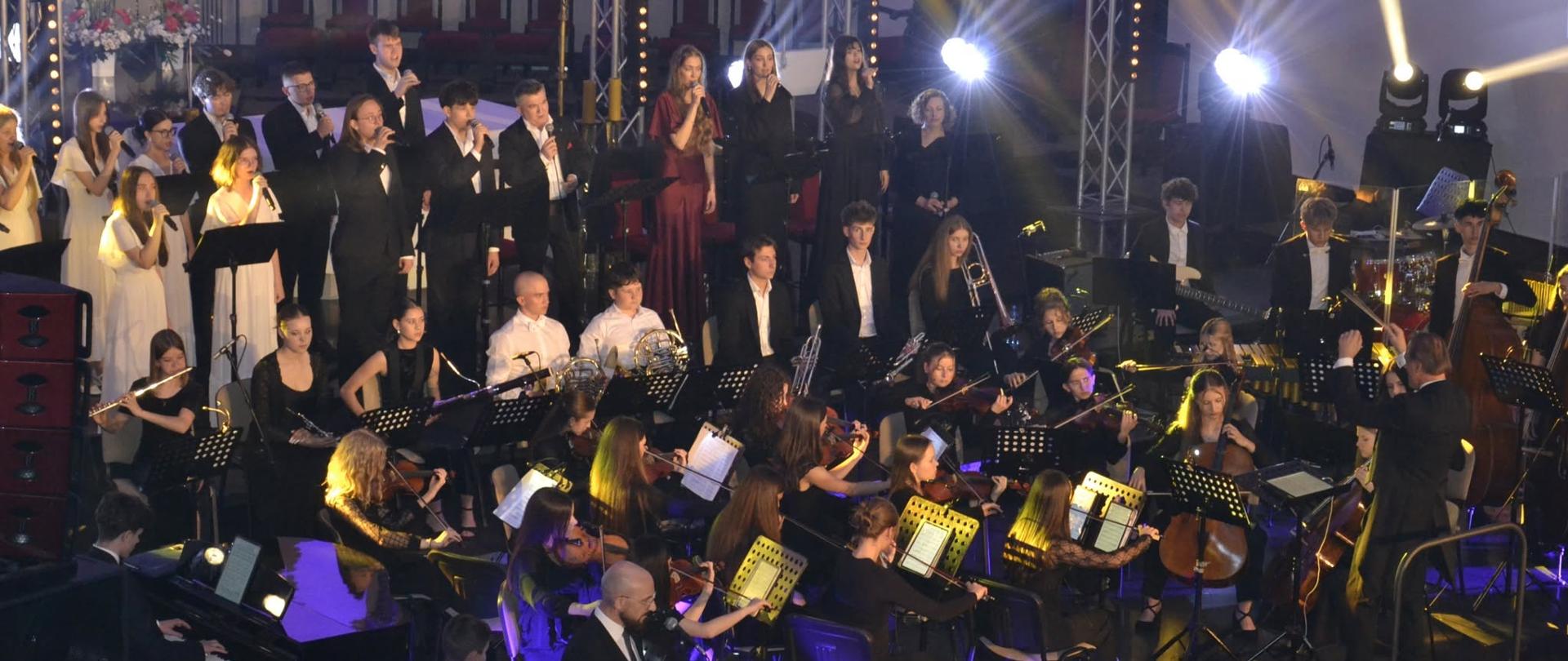 Orkiestra symfoniczna, chór i soliści PSM w Mielcu podczas wykonania „Missa Veni Sancte Spiritus" w Parafii pw. Ducha Świętego w Mielcu, oświetleni kolorowymi reflektorami scenicznymi.