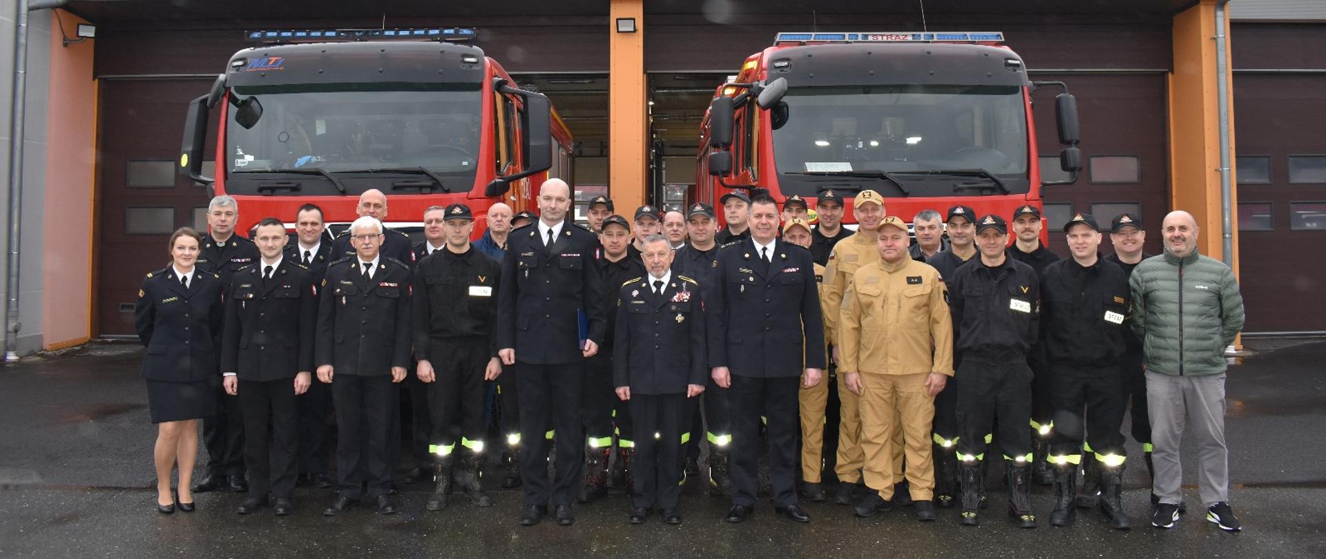 Na fotografii widać funkcjonariuszy Państwowej Straży Pożarnej na tle pojazdów pożarniczych i Jednostki Ratowniczo-Gaśniczej PSP w Jelczu-Laskowicach.