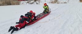 Na zdjęciu znajduje się grupa strażaków. Na pierwszym planie widać strażaka który zabezpieczony jest w ubranie niezatapialne oraz kamizelkę ratowniczą wraz z podpiętą linką asekuracyjną i trzyma strażaka pozorującego osobę poszkodowaną. Drugi strażak, zabezpieczony w kamizelkę ratowniczą wraz z podpiętą linką asekuracyjną, znajduje się na końcu sań lodowych stanowiąc przeciwwagę. Strażacy znajdujący się na brzegu stawu, ciągną liny podpięte pod sanie, w ten sposób przeciągając je po lodzie w kierunku brzegu.