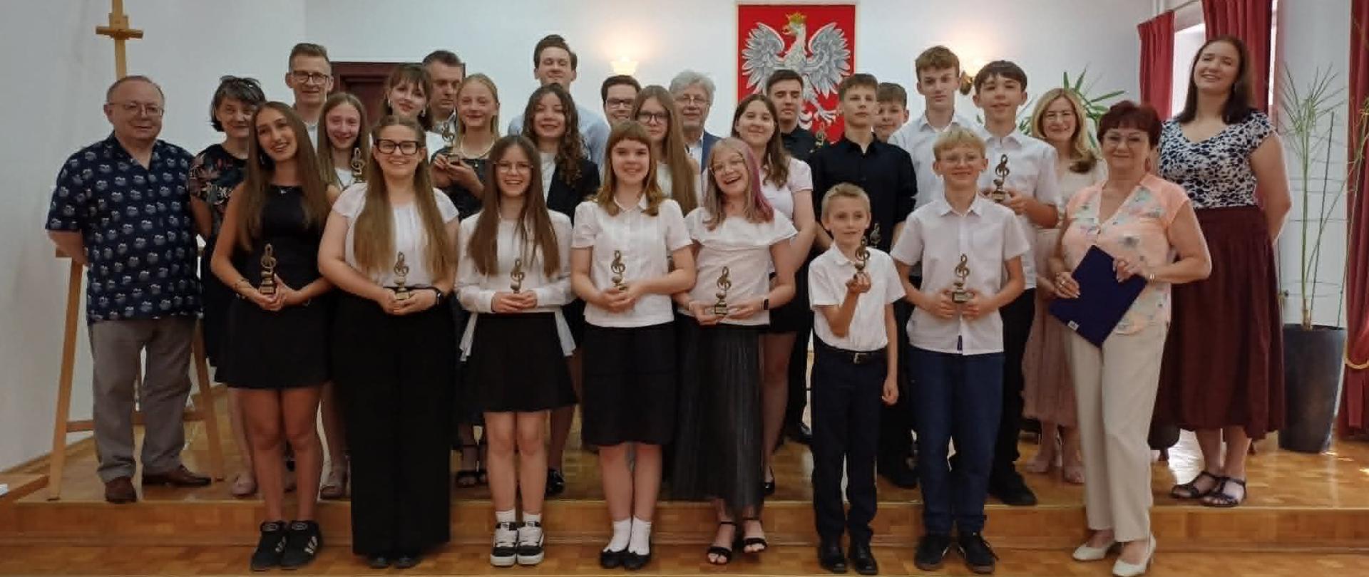 Zdjęcie przedstawia grupę absolwentów Państwowej Szkoły Muzycznej I stopnia w Bełchatowie wraz z nauczycielami oraz pracownikami szkoły stojących na scenie sali koncertowej.