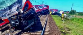 Czerwono-biały pociąg pasażerski stoi na torach po zderzeniu z ciężarówką przewożącą ładunek ponadgabarytowy. Wokół widoczne są służby ratunkowe, wozy strażackie.