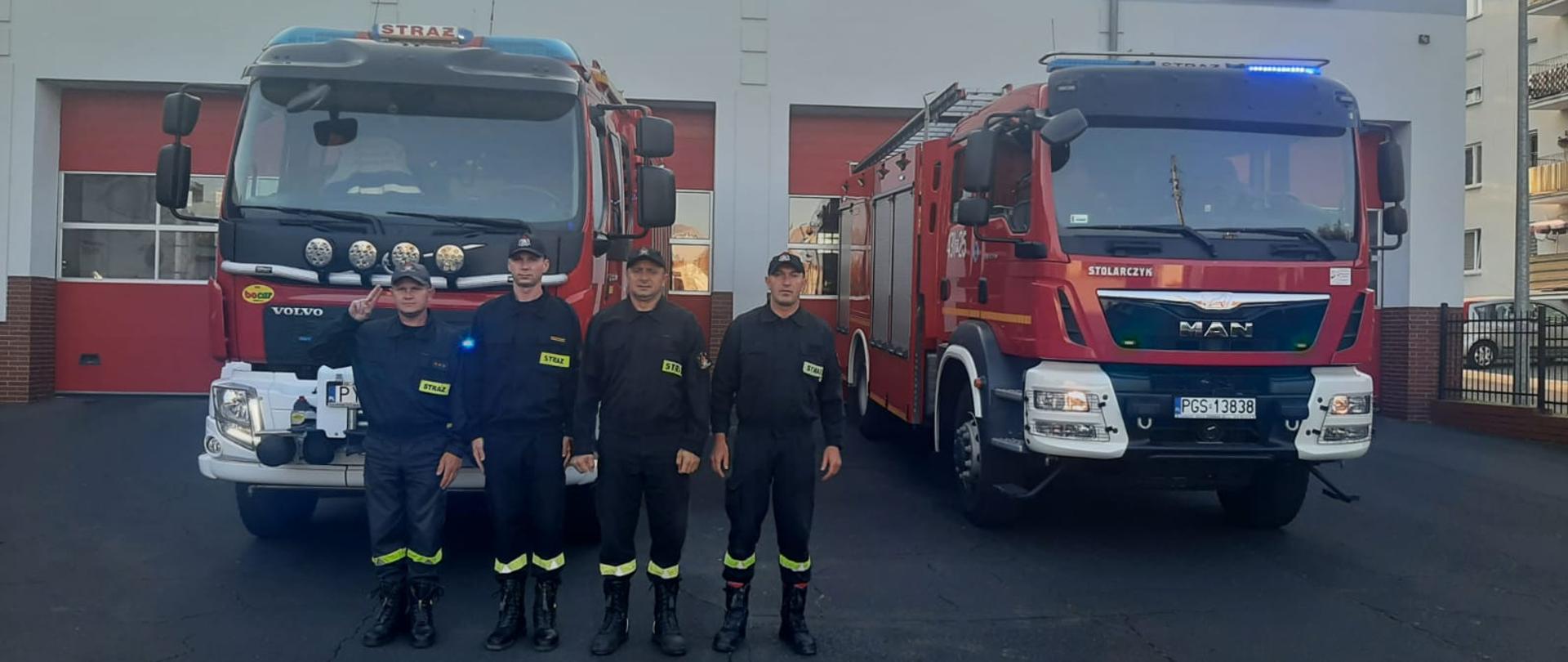 Strażacy stoją w szeregu w postawie na baczność przed wozami strażackimi. Wozy mają włączone sygnały błyskowe. Jeden ze strażaków oddaje honor poprzez salutowanie.
