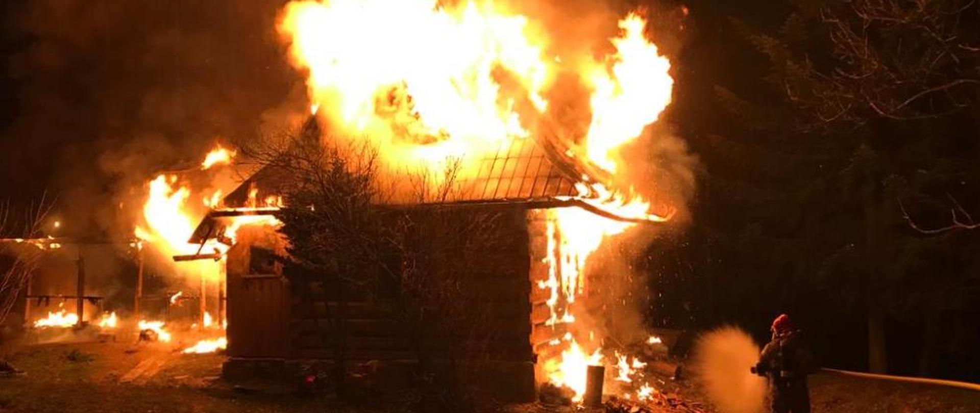 Zdjęcie przedstawia pożar budynku w Zakopanem na ul. Kotelnica. Na zdjęciu widać dom objęty w całości pożarem, po prawej stronie strażak przygotowujący się do gaszenia pożaru. Tło ciemne z uwagi na wieczorową porę zdarzenia.
