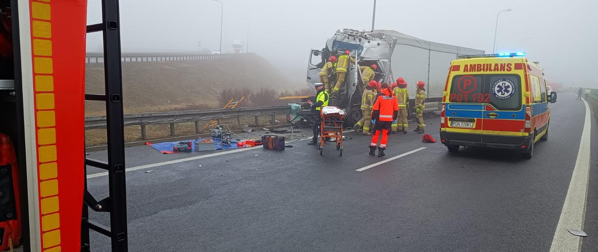 Na zdjęciu widać jak strażacy prowadzą czynności zmierzające do uwolnienia zakleszczonego kierowcy TIRa przy użyciu narzędzi hydraulicznych. Obok uszkodzonego pojazdu znajduje się karetka pogotowia. Jest duża mgła.