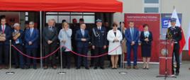 Na zdjęciu widoczne osoby na tle garażu budynku Straży pożarnej. Osoby stoją pod czerwonym namiotem. Jeden z funkcjonariuszy przemawia. Za nimi widoczne flagi biało – czerwone i brama garażowa.