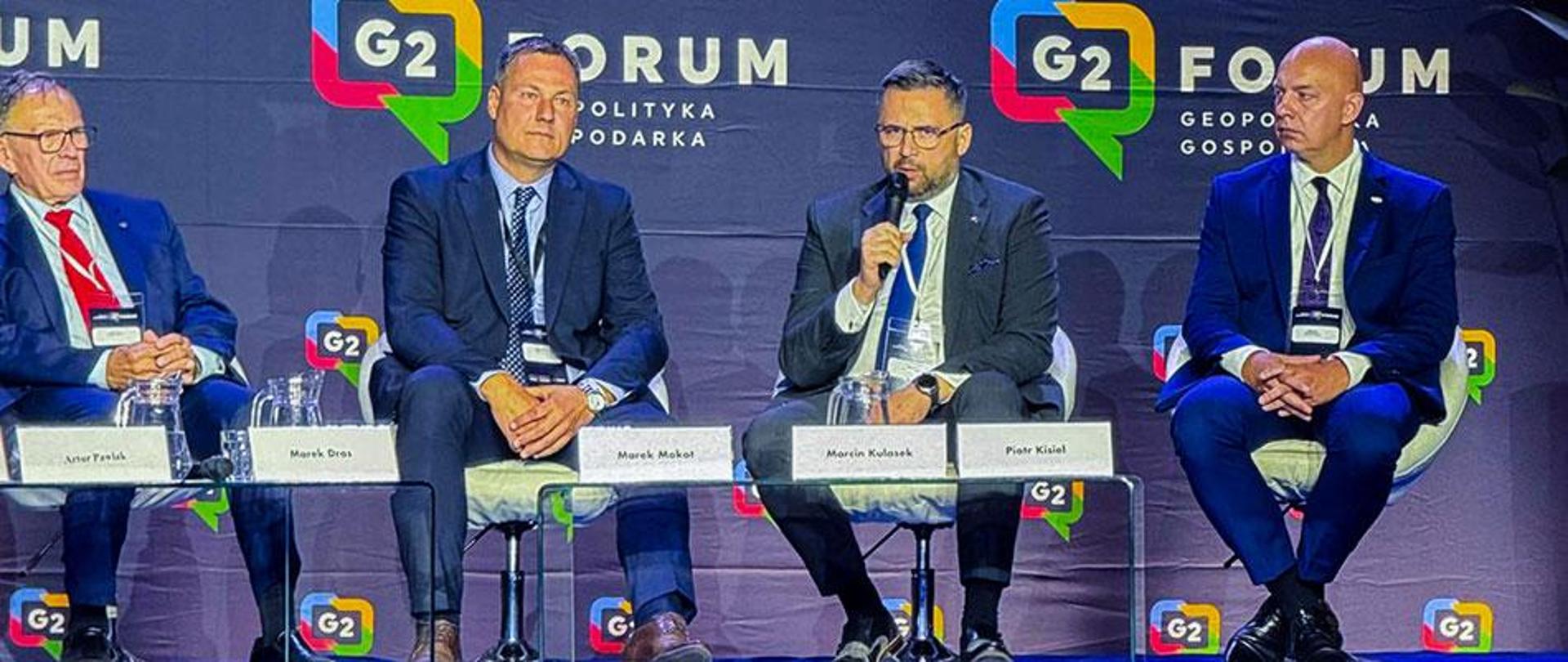 Na zdjęciu siedzi czterech mężczyzn. Przed nimi stoją stoliki z imiennymi koziołkami. W tle ścianka z logotypami Forum G2. 