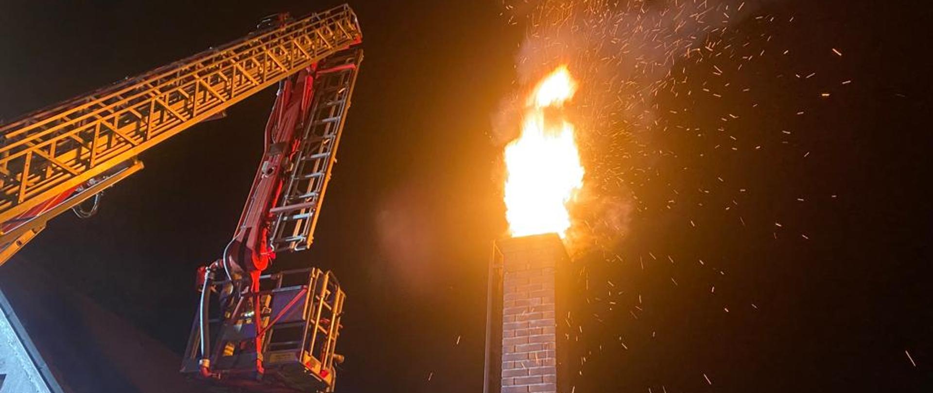 Noc. Z komina bucha ogień oraz iskry. Z lewej strony kosz i drabina mechaniczna.