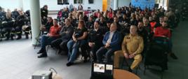 Strażacy Państwowej Straży Pożarnej druhowie ochotniczych straży pożarnych oraz zaproszeni goście siedzą na krzesłach przed nimi ustawiony jest na stoliku rzutnik oraz laptop.