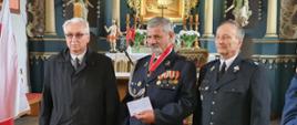 Prezesa Ochotniczej Straży Pożarnej w Gwieździnie druha Antoniego Wiśniewskiego otrzymującego odznaczenie oraz dokonujących wręczenia dh. Grzegorza Pladzyk- sekretarza Zarządu Oddziału Wojewódzkiego ZOSP RP województwa pomorskiego i dh. Tadeusza Lisowskiego - Prezes Zarządu Oddziału Powiatowego ZOSP RP w Człuchowie. W tle zabytkowy ołtarz kościoła w Gwieździnie.