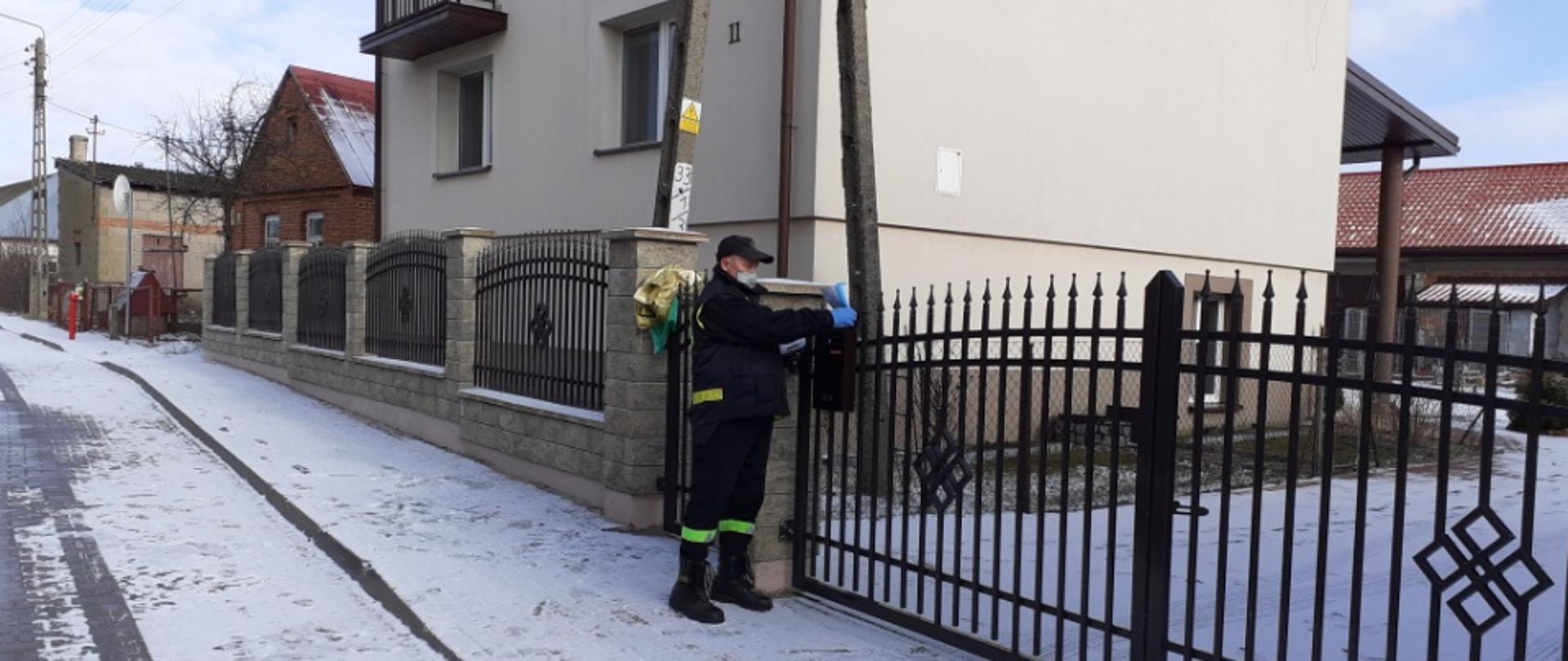 Strażak OSP wkładający ulotkę do skrzynki na listy przy bramie wjazdowej na posesję.
