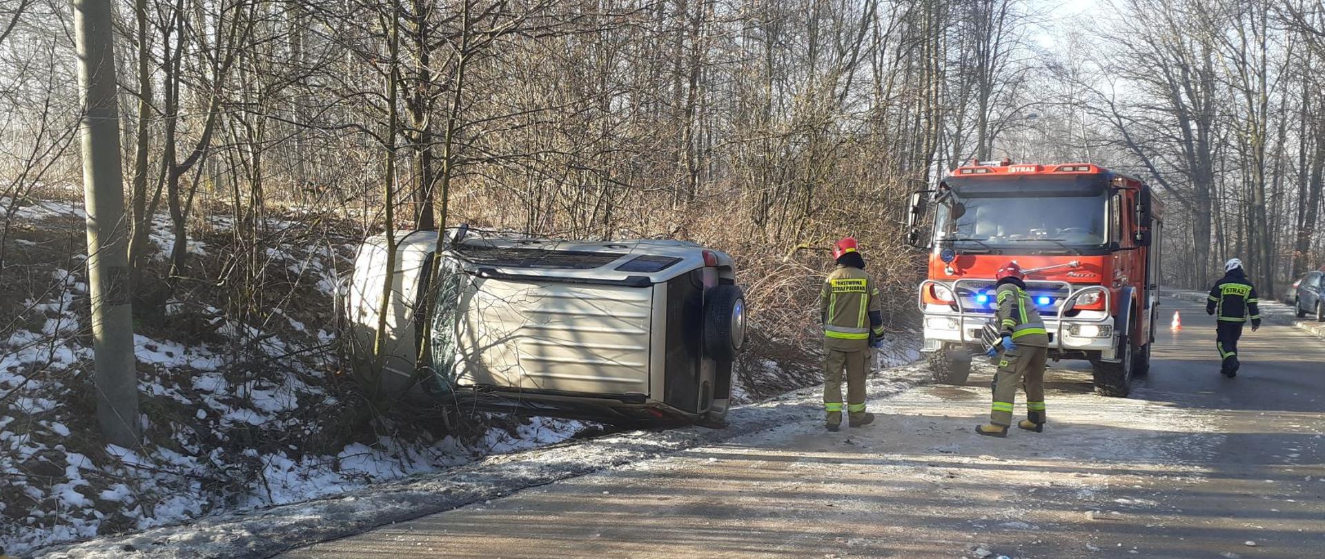 Na zdjęciu widoczny samochód leżący na lewym boku. Strażacy zwiększają szorstkość nawierzchni jezdni w celu bezpiecznego prowadzenia działań. Średni samochód ratowniczo- gaśniczy zabezpiecza miejsce zdarzenia stojąc na niebieskich światłach błyskowych.