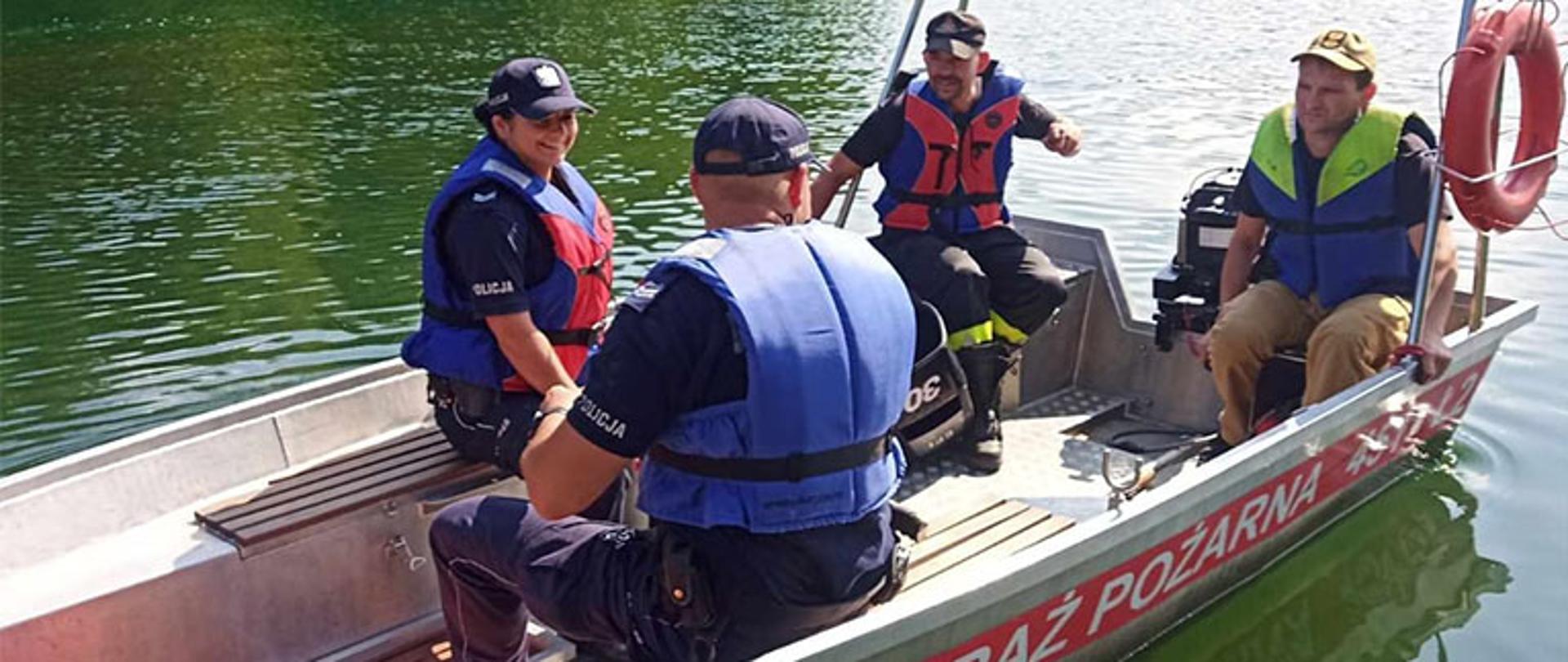 na terenie akwenu wodnego cztery osoby siedzą w aluminiowej łodzi z napisem państwowa straż pożarna na lewej burcie, wszyscy maja założone kamizelki ratunkowe, w tle zalesiony brzeg zbiornika wodnego, pogoda słoneczna, niebo bezchmurne
