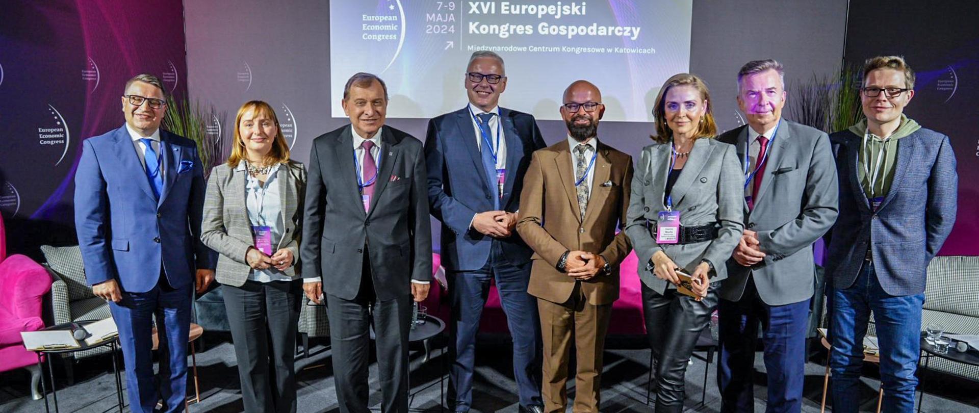 Pod fioletową ścianą z napisem XVI Europejski Kongres Gospodarczy stoi w szeregu osiem osób, minister Wieczorek drugi od prawej.