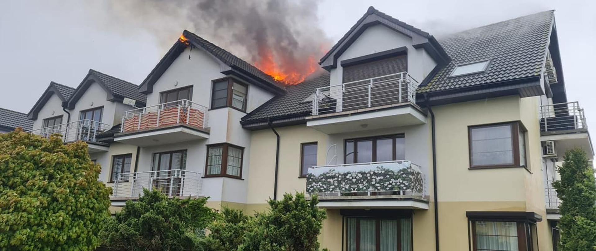 Zdjęcie przedstawia budynek wielorodzinny z widocznymi płomieniami na dachu.