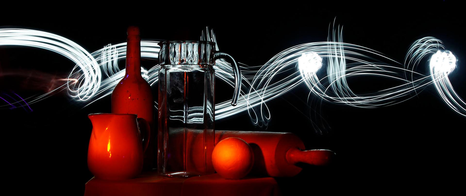 Fotografia martwej natury na czarnym tle. Wałek do ciasta. szklany dzbanek, butelka, dzbanek emaliowany oświetlone są ciepłym światłem. Za naczyniami smuga światła w chaotycznym ruchu.