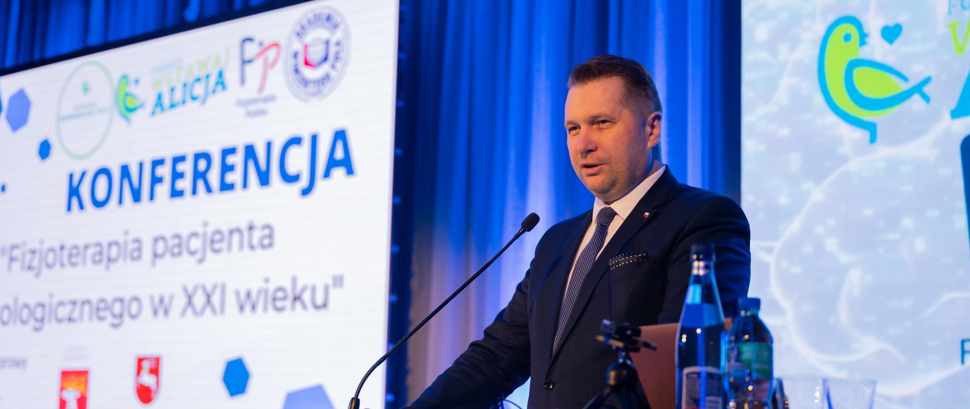 Przy mównicy stoi minister Czarnek i mówi do mikrofonu, za nim na ścianie napis Konferencja „Fizjoterapia pacjenta neurologicznego w XXI wieku”.
