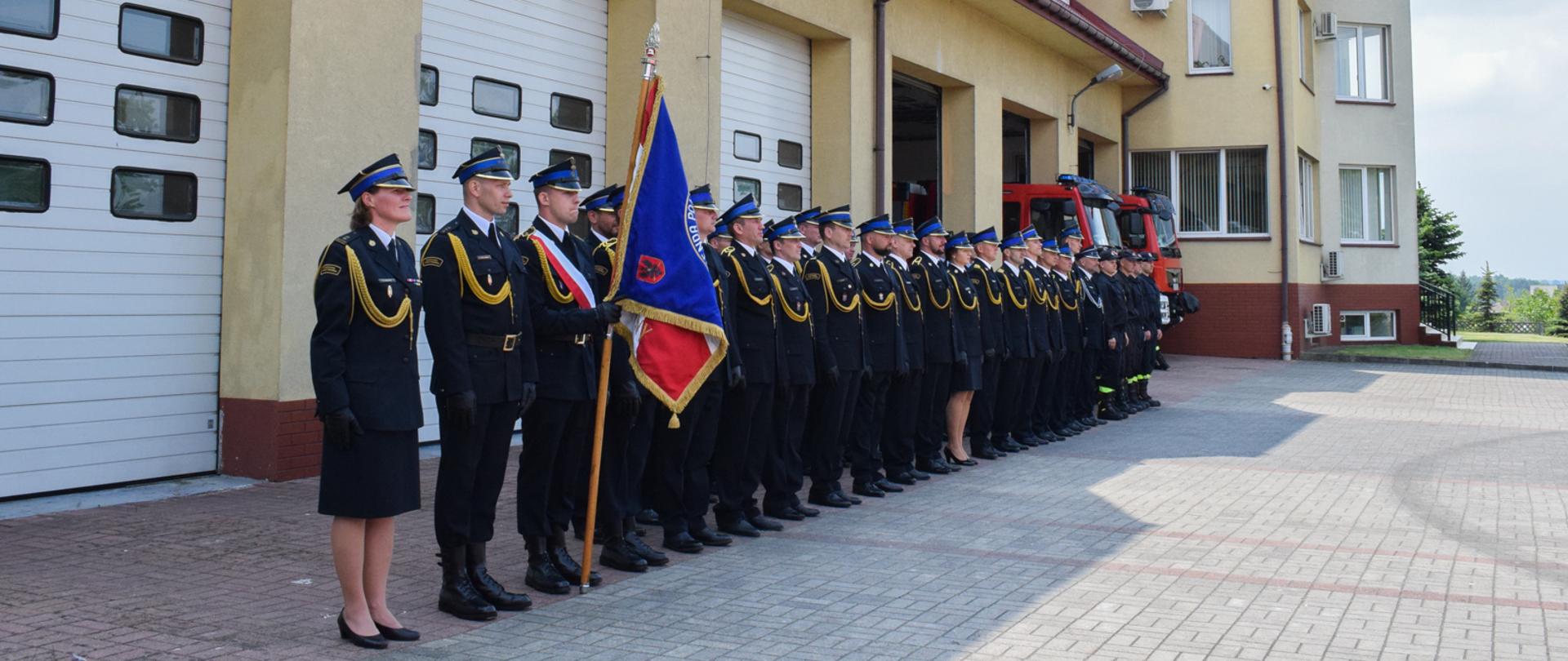 strażacy wraz z pocztem sztandarowym stoją na zbiórce przed garażami komendy