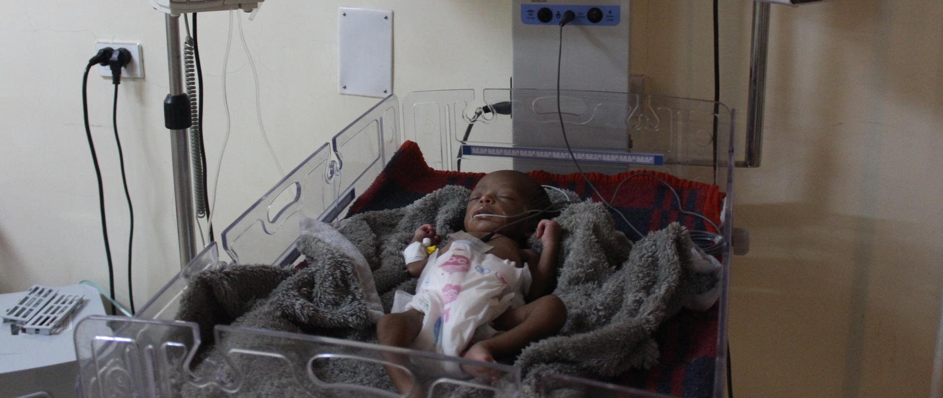Niemowlę leży podpięte do sprzętu medycznego w za dużej pieluszce, na kocyku