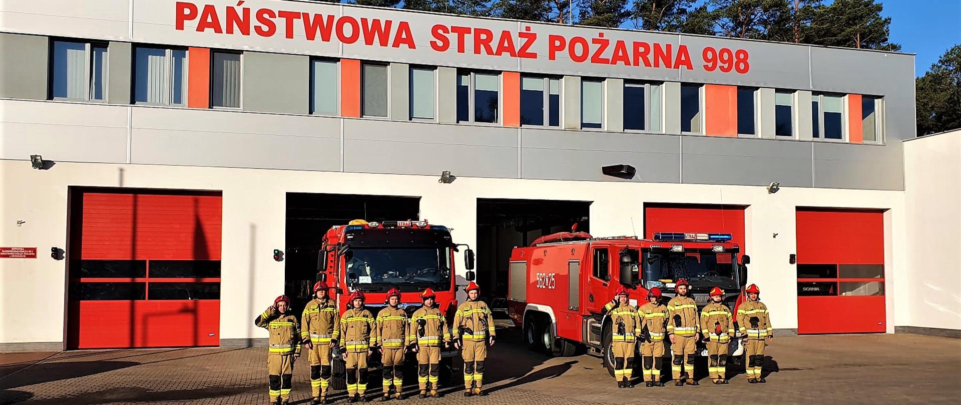 Zmiana służby w JRG 2. Samochody bojowe przed garażami. Strażacy zmian służbowych w mundurach bojowych i hełmach stoją w szeregu. Dowódcy zmian oddają honor.