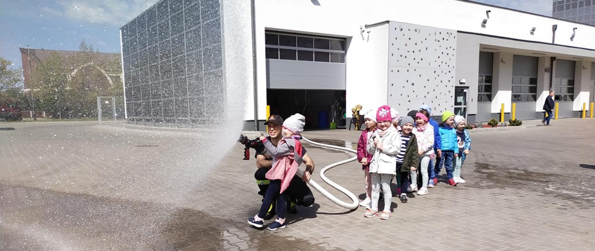 Dzieci ustawione na tle Komendy leją wodę za pomocą węża strażackiego.