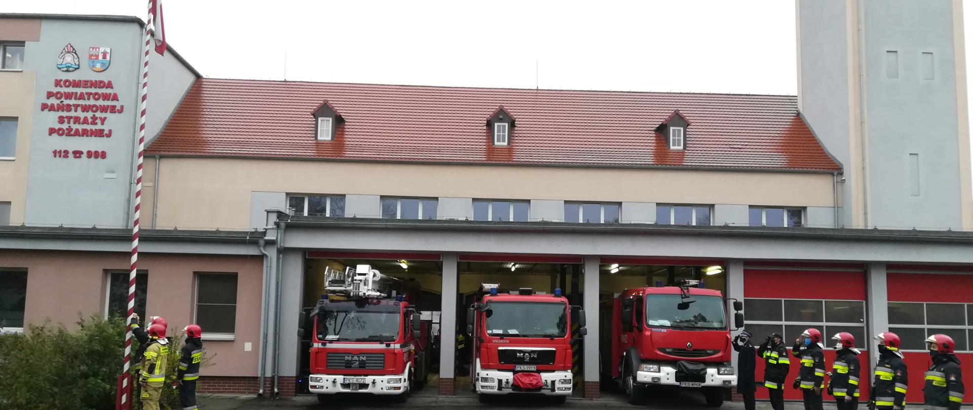 Narodowe Święto Niepodległości. Na zdjęciu strażacy zawieszający flagę Polski na maszt. Strażacy ustawieni w szyku. W tle widać samochody pożarnicze oraz budynek Komendy.