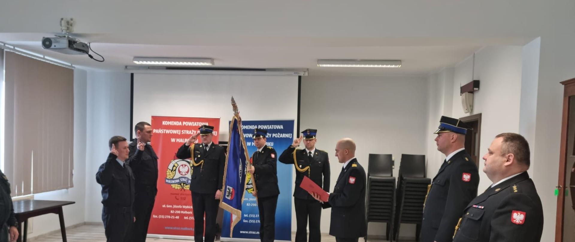 Strażacy stoją z oddanymi honorami i sztandarem podczas odczytywania roty ślubowania.