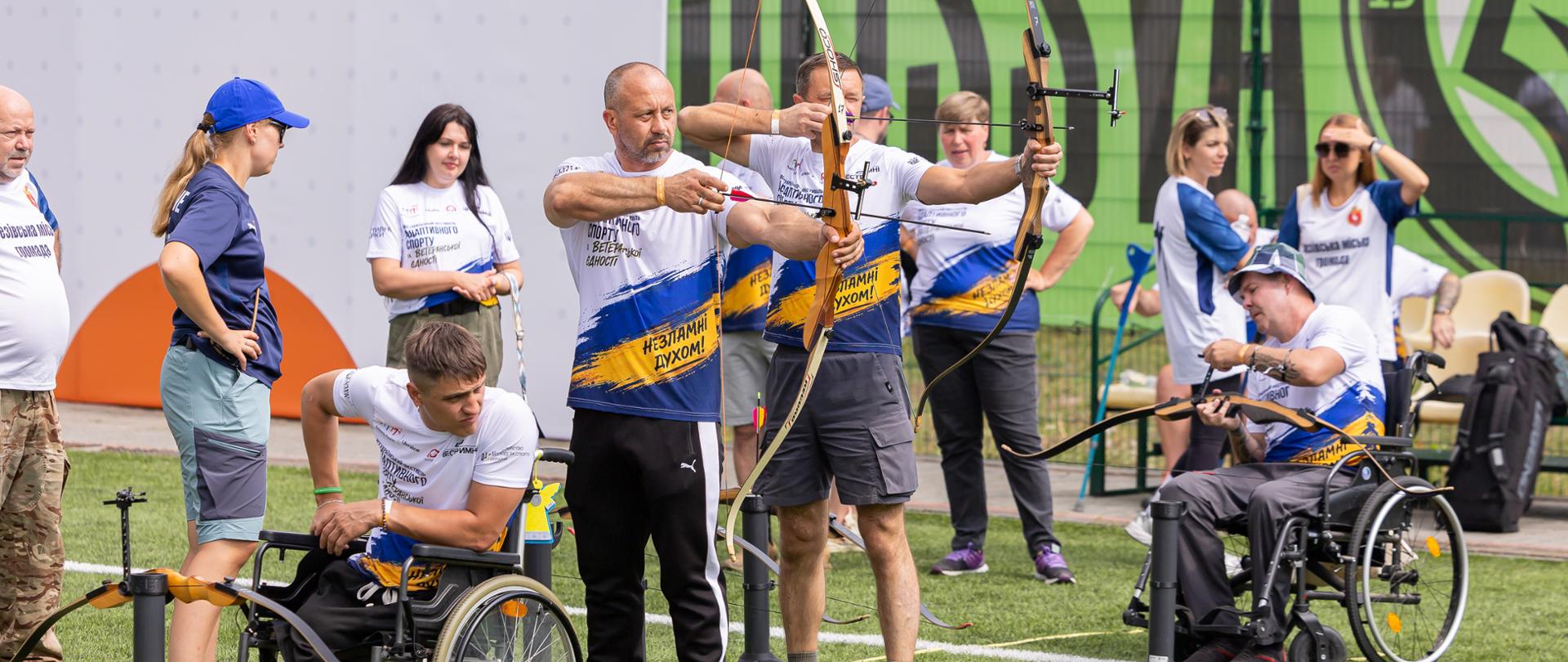 Grupa ludzi z łukami, dwóch mężczyzn na wózkach inwalidzkich, wszyscy mają na sobie sportowe koszulki