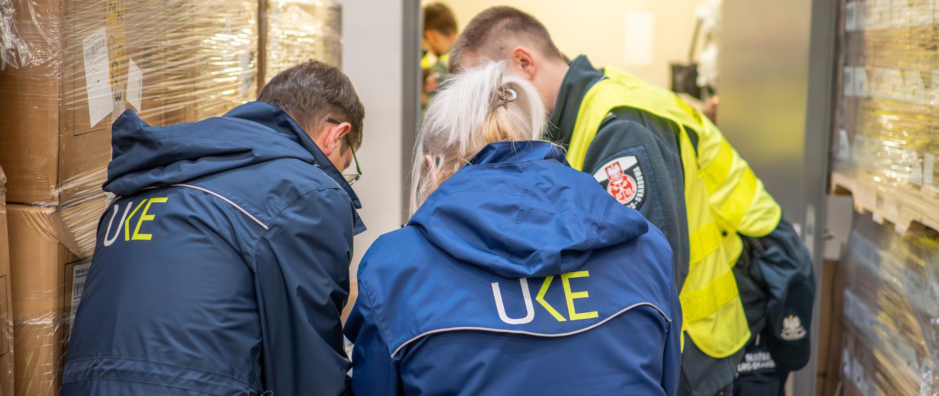 Trzy osoby w odzieży służbowej pochylają się nad stołem w magazynie, gdzie w tle znajdują się palety owinięte folią. Na plecach dwóch osób widoczny jest napis „UKE”, a obok stoi osoba w odblaskowej kamizelce