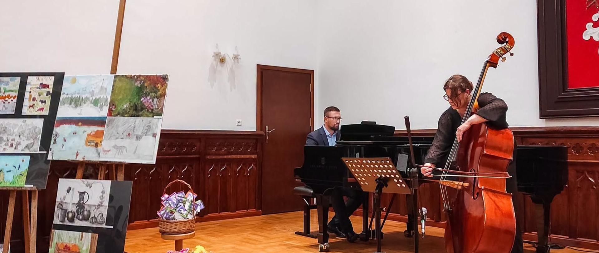 Nauczycielka gra na kontrabasie, a nauczyciel akompaniuje na fortepianie na scenie Sali Królewskiej; obok ekspozycja prac plastycznych uczniów studia art.GOYA.