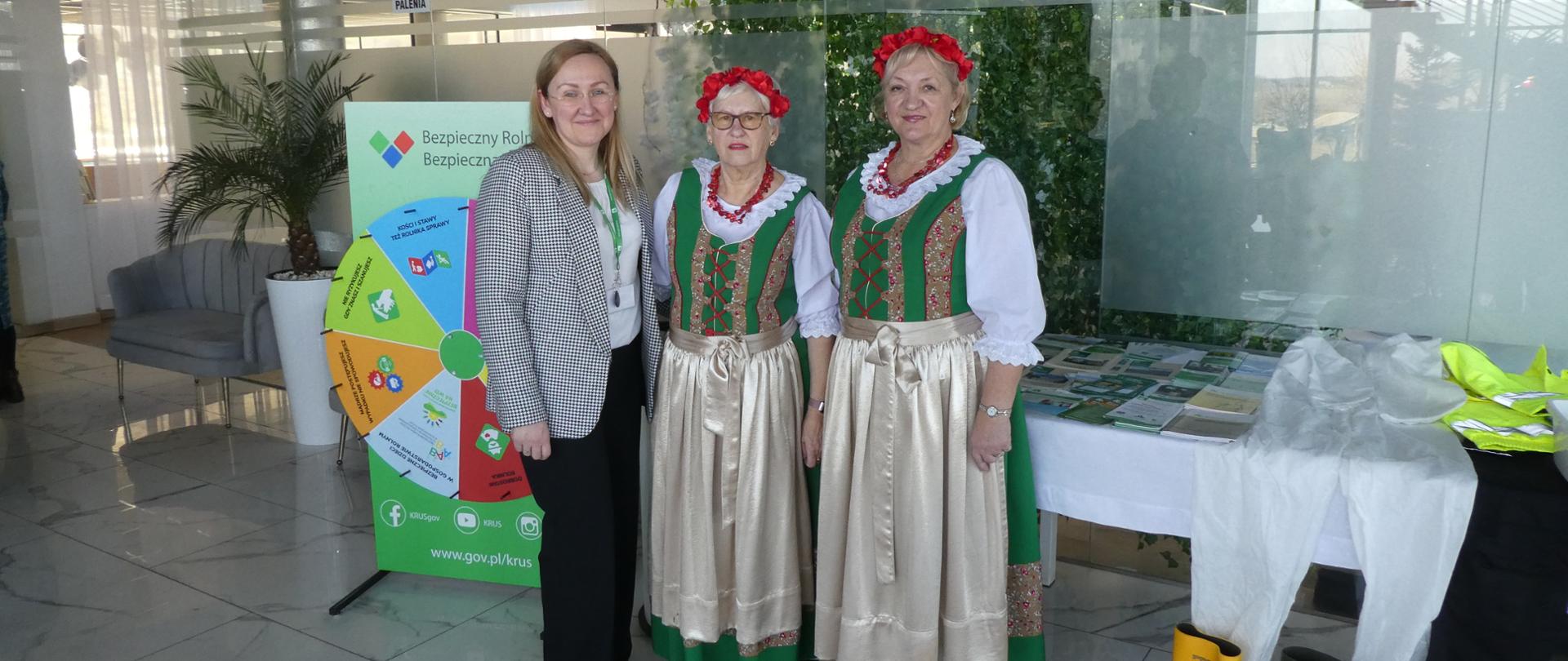 Trzy kobiety (dwie w strojach ludowych) stoją w hallu przy stole, na którym leżą materiały informacyjne KRUS, obok na stole leży rozłożony kombinezon i obuwie ochronne do prac rolniczvch. Z tyłu po lewej stoi gra prewencyjna "Koło Fortuny".