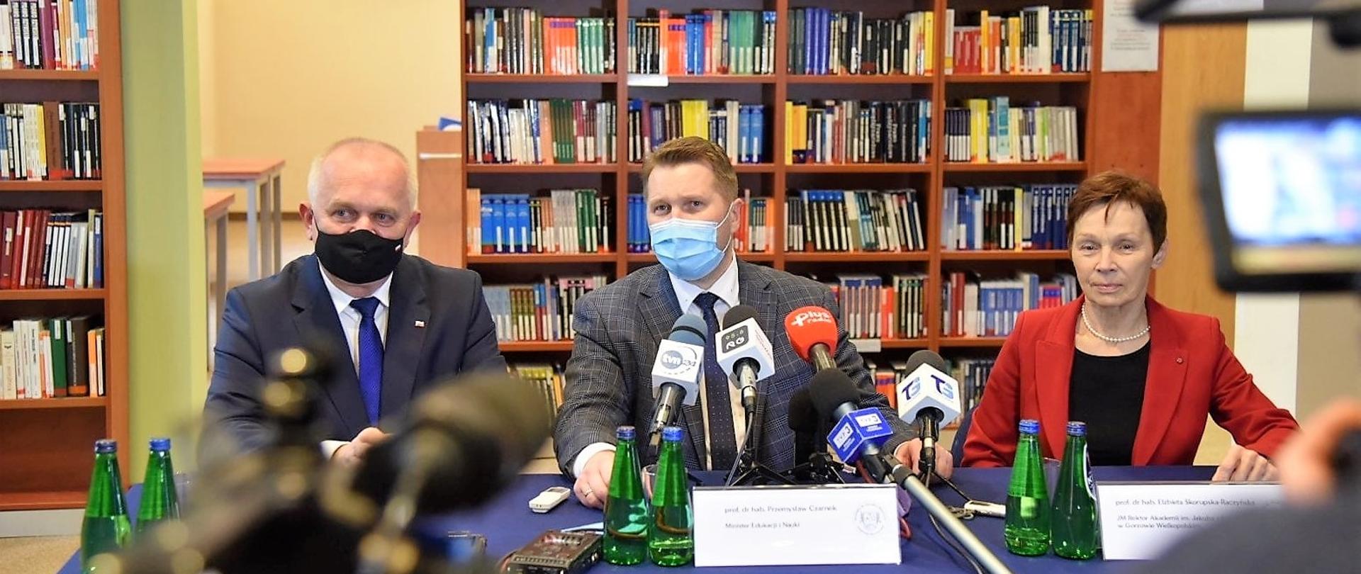 W czasie konferencji prasowej w bibliotece Akademii (regały z książkami w tle) przy stole siedzi wojewoda, minister i pani rektor. Zdjęcie zrobione zza kamer, ich fragmenty widać u dołu i po prawej stronie 
