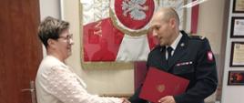 Zdjęcie przedstawia jubilatkę oraz p.o. komendanta na tle sztandaru KP PSP podczas wręczania nagrody jubileuszowej