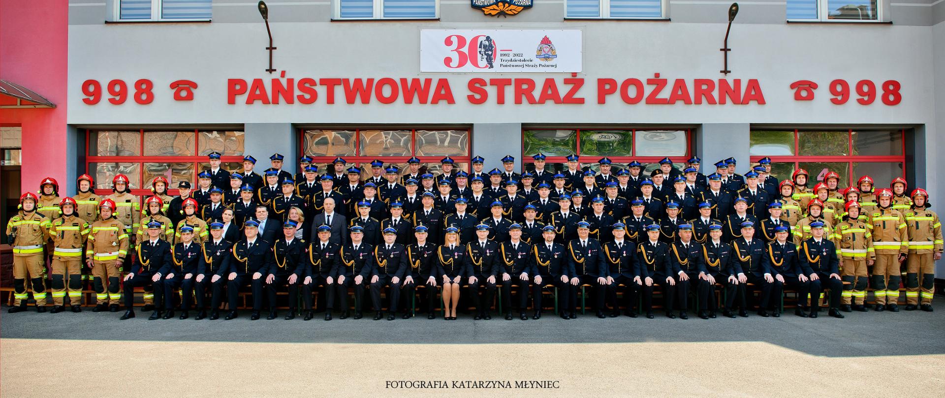 Przed budynkiem strażnicy strażacy i pracownicy cywilni pozują do fotografii. W środkowej części fotografii znajdują się strażacy w umundurowaniu wyjściowym, a po prawej i lewej stronie strażacy w umundurowaniu bojowym. Wśród strażaków znajdują się pracownicy w ubiorach cywilnych. Na elewacji budynku znajduje się napis "998 PAŃSTWOWA STRAŻ POŻARNA 998" widoczne również logo Państwowej Straży Pożarnej oraz baner przedstawiający informację o jubileuszu trzydziestolecia PSP. 
