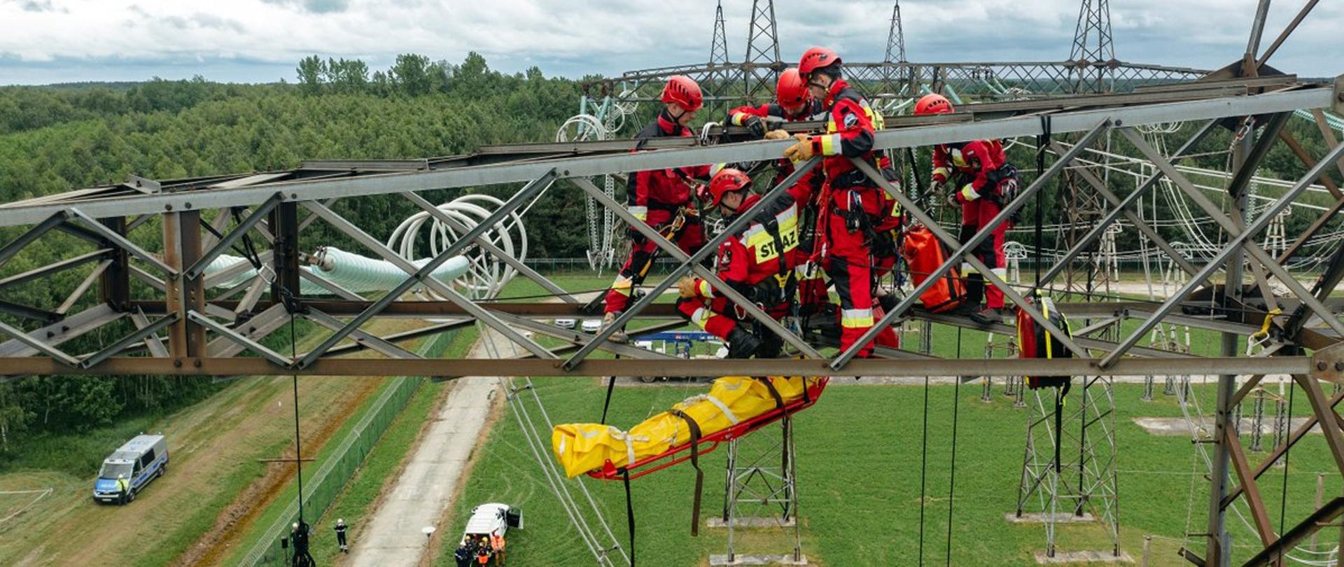 Zdjęcie zrobione na zewnątrz, podczas ćwiczeń strażaków na słupie wysokiego napięcia. Strażacy ze specjalistycznej grupy ratownictwa wysokościowego realizują epizod ćwiczeń.