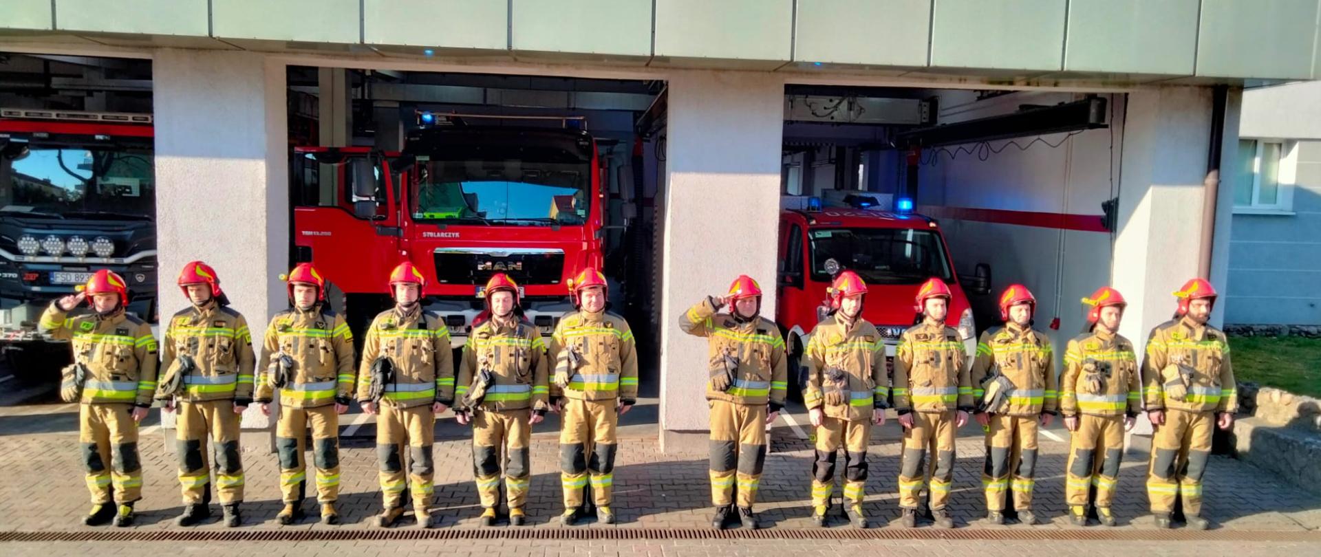 Fotografia przedstawia moment uczczenia minutą ciszy poległych strażaków Ukrainy. Dwie zmiany stoją w jednym rzędzie przed garażem JRG PSP w Strzelcach Krajeńskich. Dowódcy zmian salutują, wszyscy ubrani w ubrania specjalne i hełmy. Jest dzień, słonecznie.
