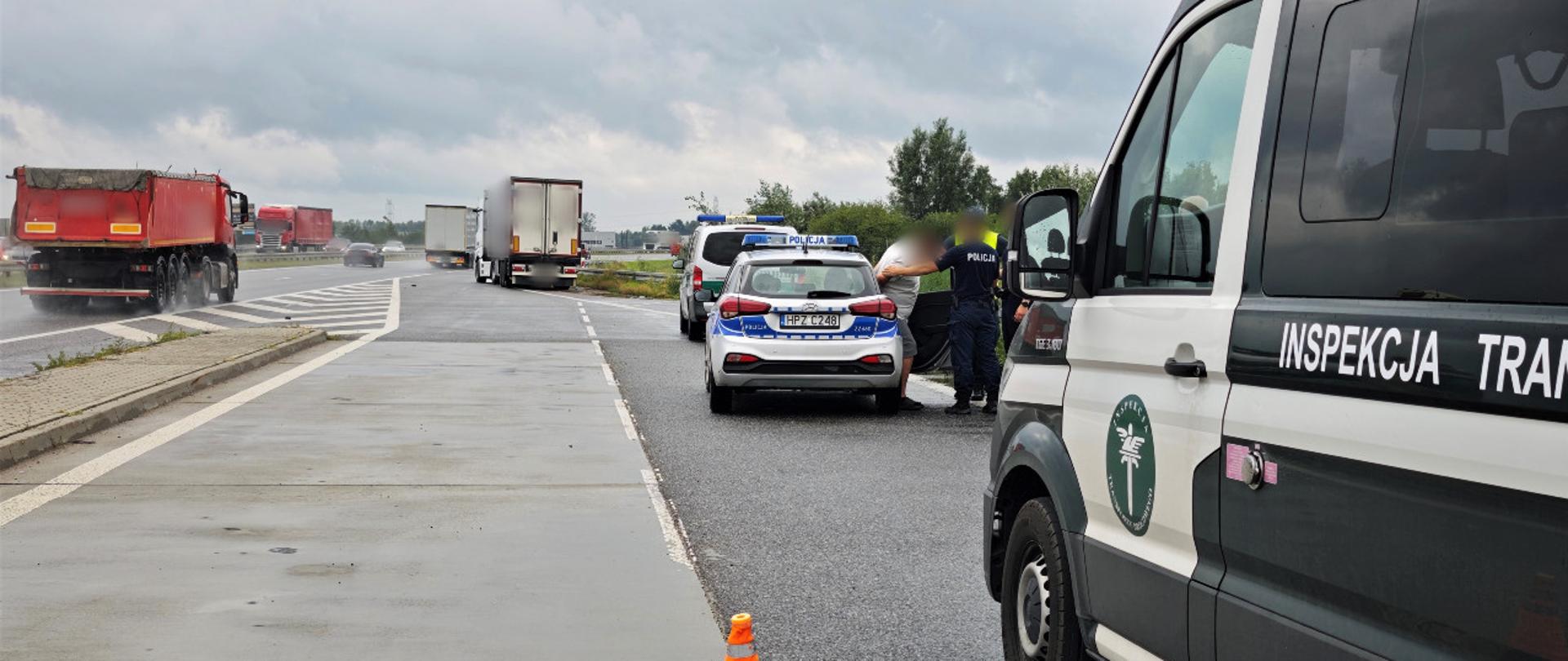 Wysiadający z radiowozu w asyście Policjantów kierowca ciężarówki. Obok stoi oznakowany radiowóz ITD. W oddali ciężarówka. 