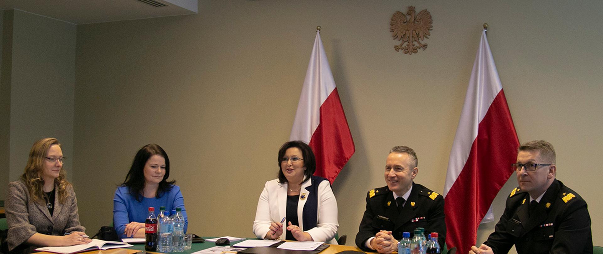 Na zdjęciu przedstawiona jest grupa osób siedząca na tle dwóch biało-czerwonych flag oraz godła Polski. Przy biurku od prawej strony siedzi nadbrygadier Krzysztof Hejduk - Zastępca Komendanta Głównego Państwowej Straży Pożarnej, dalej nadbrygadier Andrzej Bartkowiak – Komendant Główny PSP, w środku Pani Małgorzata Zdrodowska – Dyrektor Zakładu Emerytalno-Rentowego MSWiA, obok Pani Aneta Marzec Naczelnik Wydziału Kadrowo-Organizacyjnego oraz po lewej Pani Justyna Borowska z Wydziału Kadrowo-Organizacyjnego.