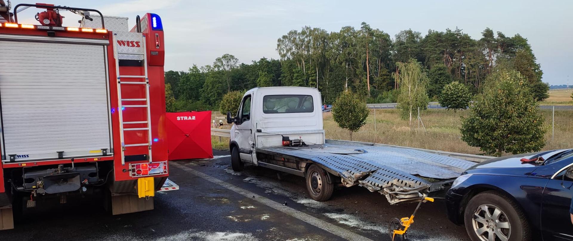 Na zdjęciu widać tył samochodu pożarniczego. Obok stoi autolaweta a za nią samochód osobowy, który był wciągany na autolawetę. Przed pojazdami widać czerwony parawan strażacki
