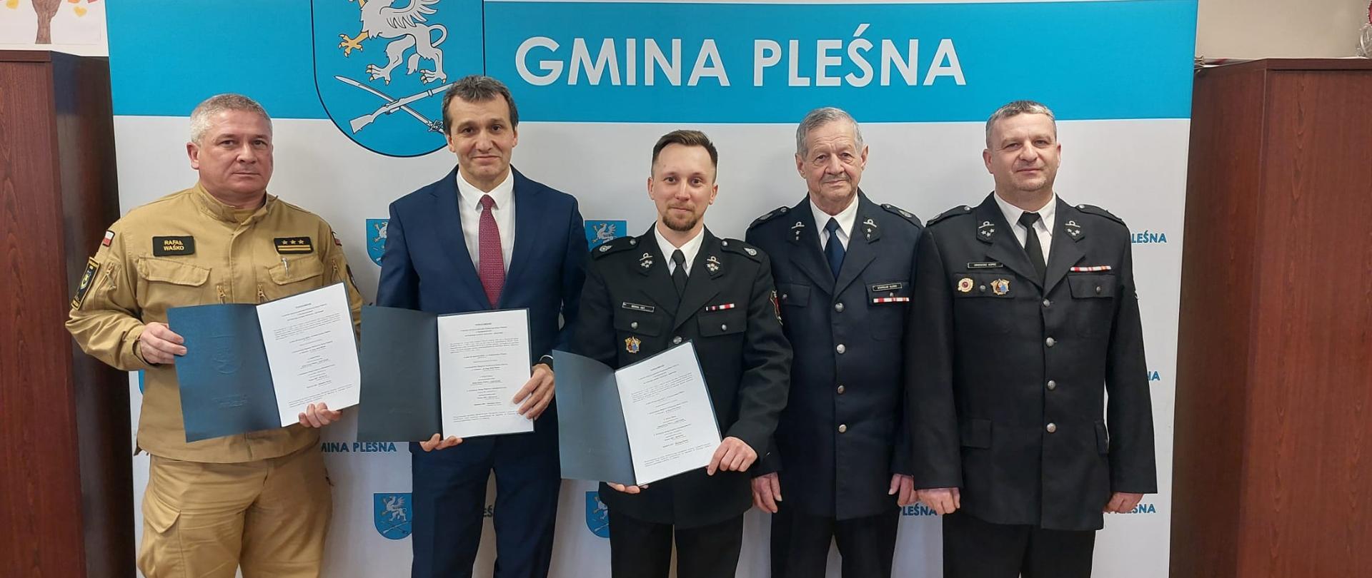 Przed niebiesko białym banerze Gminy Pleśna stoją w rzędzie strażacy ubrani w mundury oraz wójt ubrany w garnitur. W rękach trzymają porozumienie.