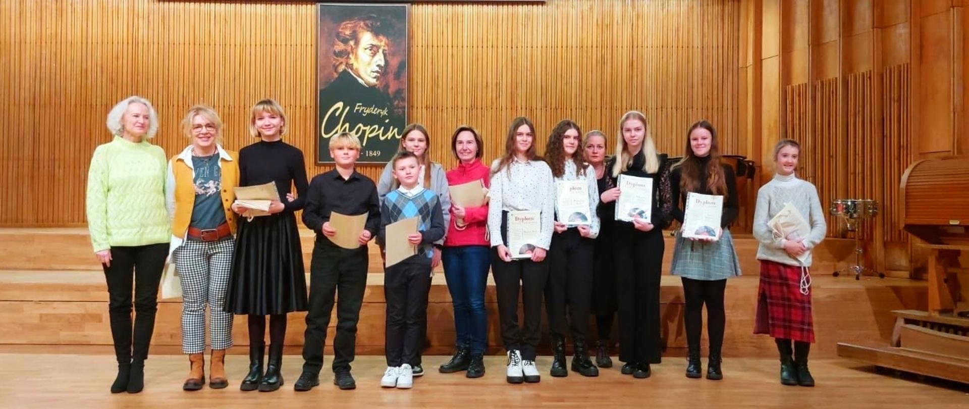 na scenie koncertowej stoją nagrodzeni uczniowie wraz z nauczycielami, w tle portret Chopina