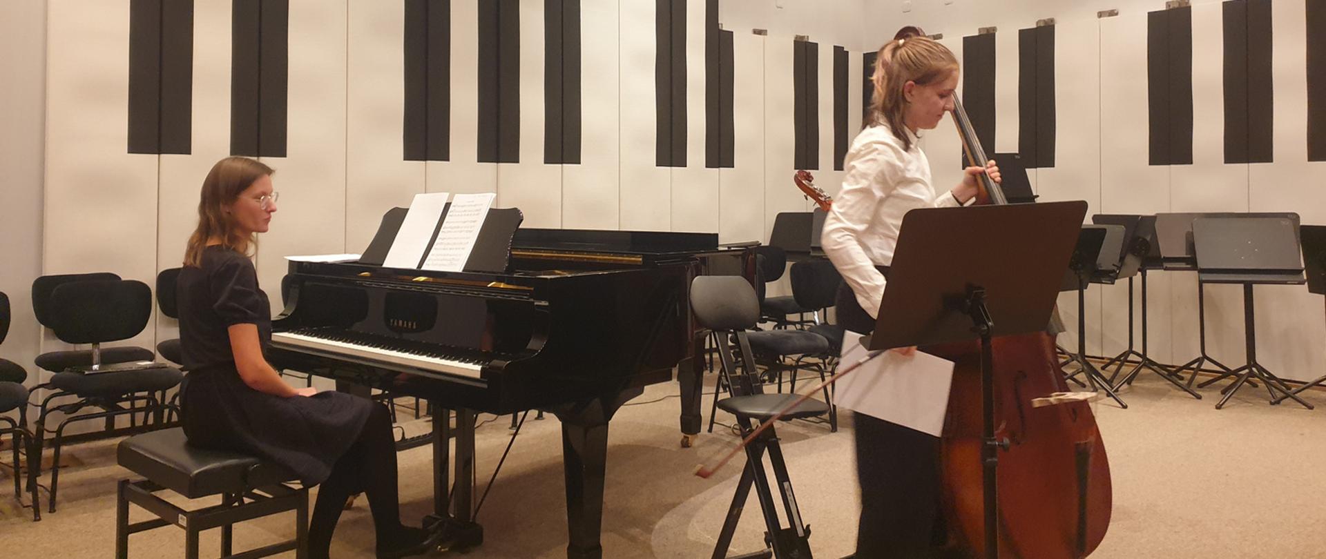 Nastolatka stoi na estradzie auli PSM z kontrabasem, w prawej ręce trzyma smyczek i buty. Przed nią stoi pulpit, za nią wysokie krzesło oraz fortepian przy którym siedzi kobieta.