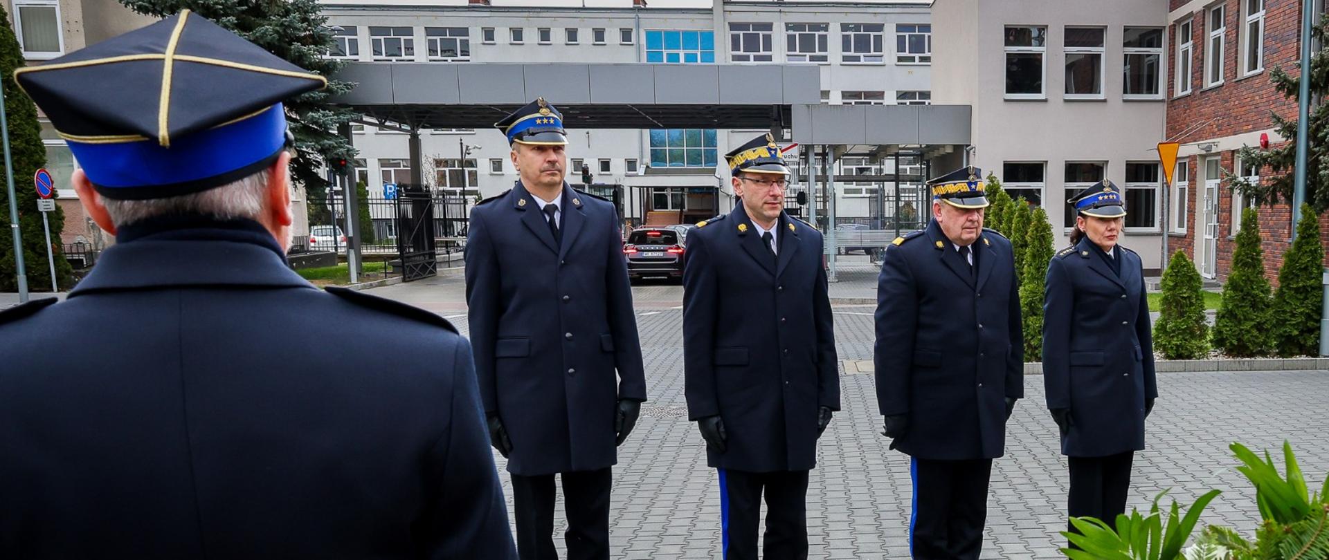 Funkcjonariusze Państwowej Straży Pożarnej stoją w skupieniu i ciszy przed pomnikiem, składając symboliczny hołd poległym. Zdjęcie ukazuje chwilę powagi i zadumy.
