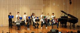 Scena w kolorze berzowym-brązowym, na której występuje zespół instrumentalny w strojach biało-czarnych, dzieci z przodu siedzą na krzesłach, dzieci z tyłu stoją