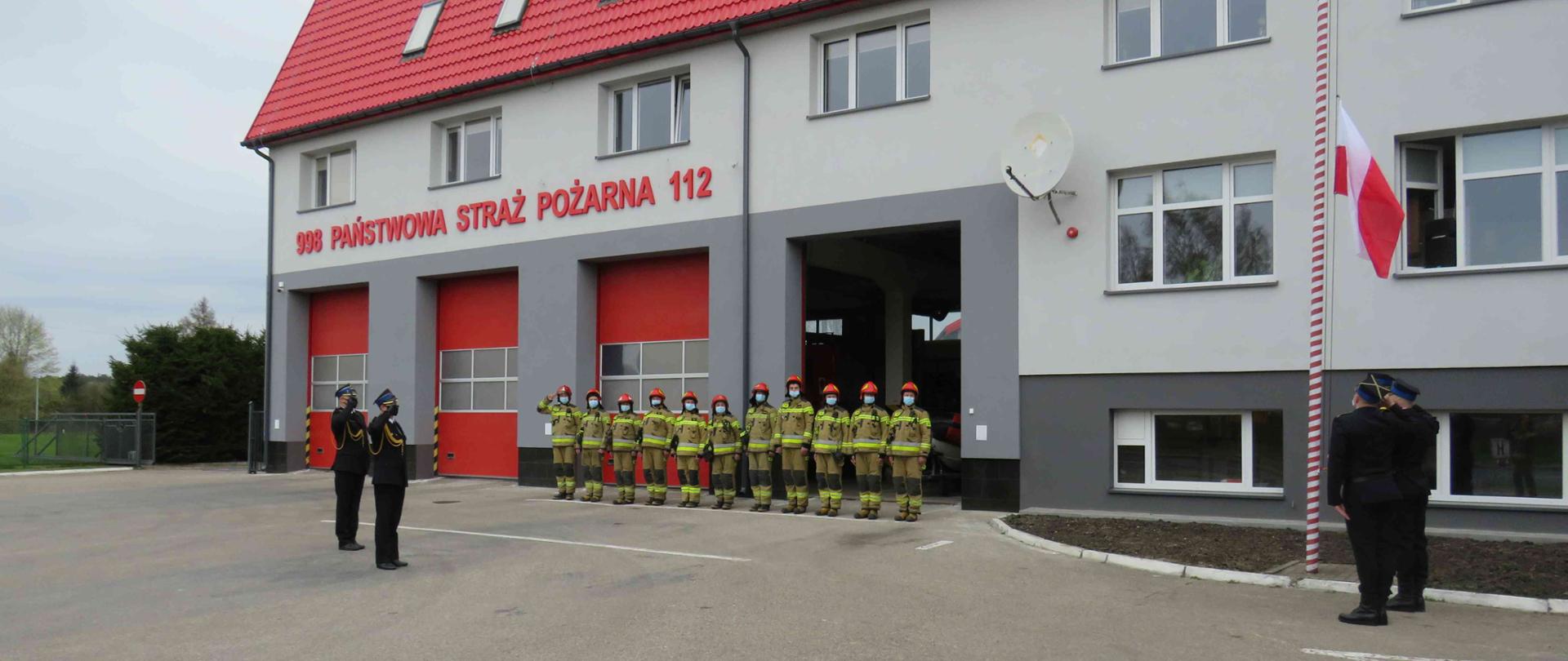 Strażacy stojący przed remizą i salutujący podczas wciągania flagi na maszt.