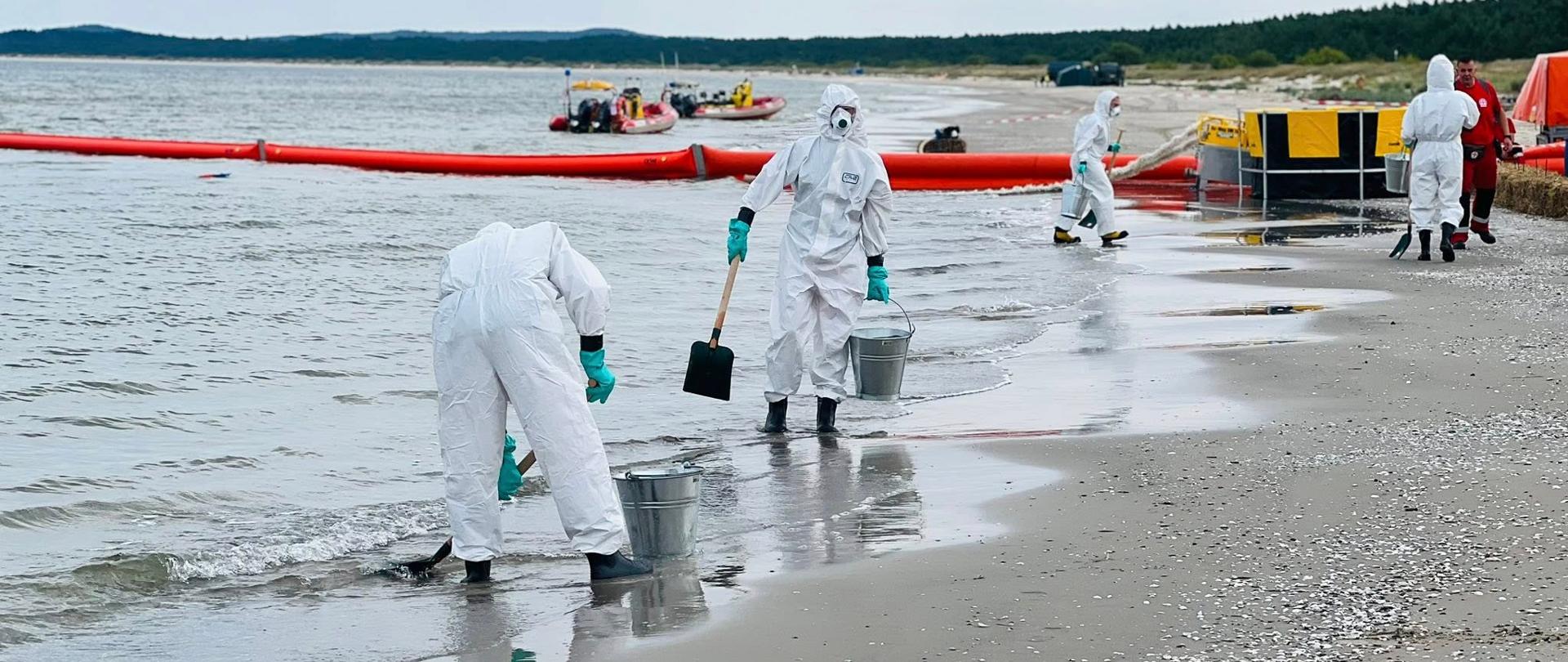 Ratownicy w białych kombinezonach ochronnych zbierają zanieczyszczenia z linii brzegowej przy pomocy wiader i łopat. W tle rozstawione zapory przeciwrozlewowe na wodzie oraz pontony ratownicze.