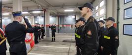 W prawej części zdjęcia strażacy stojący w dwuszeregu, na środku ślubujący w lewej części Komendant i inni strażacy. W lewej części zdjęcia rozwinięty sztandar. Zdjęcie wykonane na garażu komendy. 